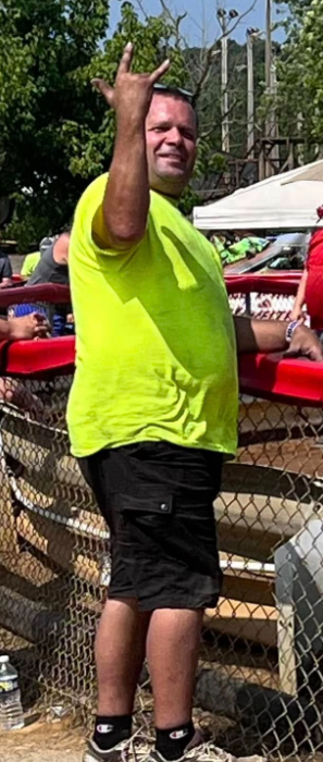 Jeff in a bright green shirt waving outdoors beside a fence.
